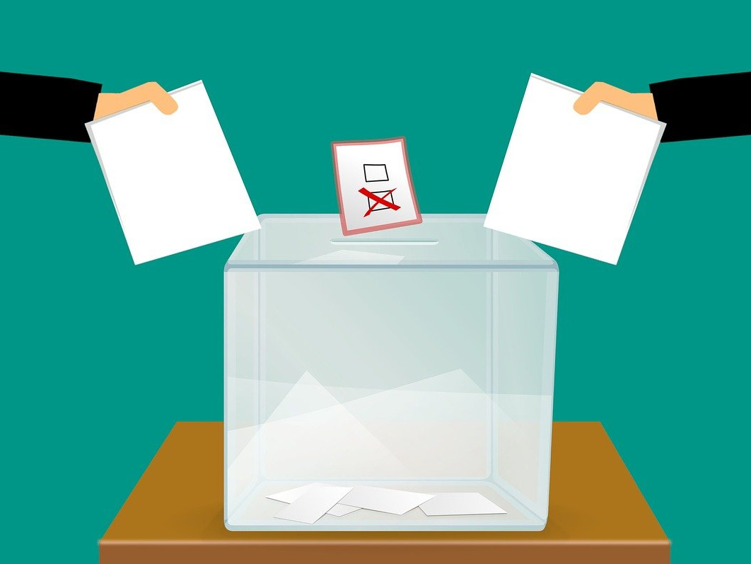Grafika przwdstawia urnę do głosowania i wyciągniete recę z kartami nad nią