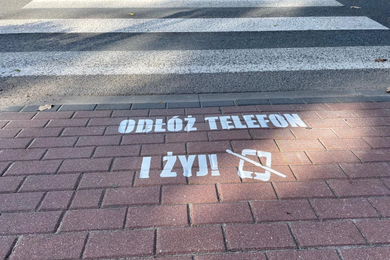 Zdjęcie przedstawia biały napis na chodniku przed przejściem dla pieszych o treści: "Odłóż telefon i żyj"